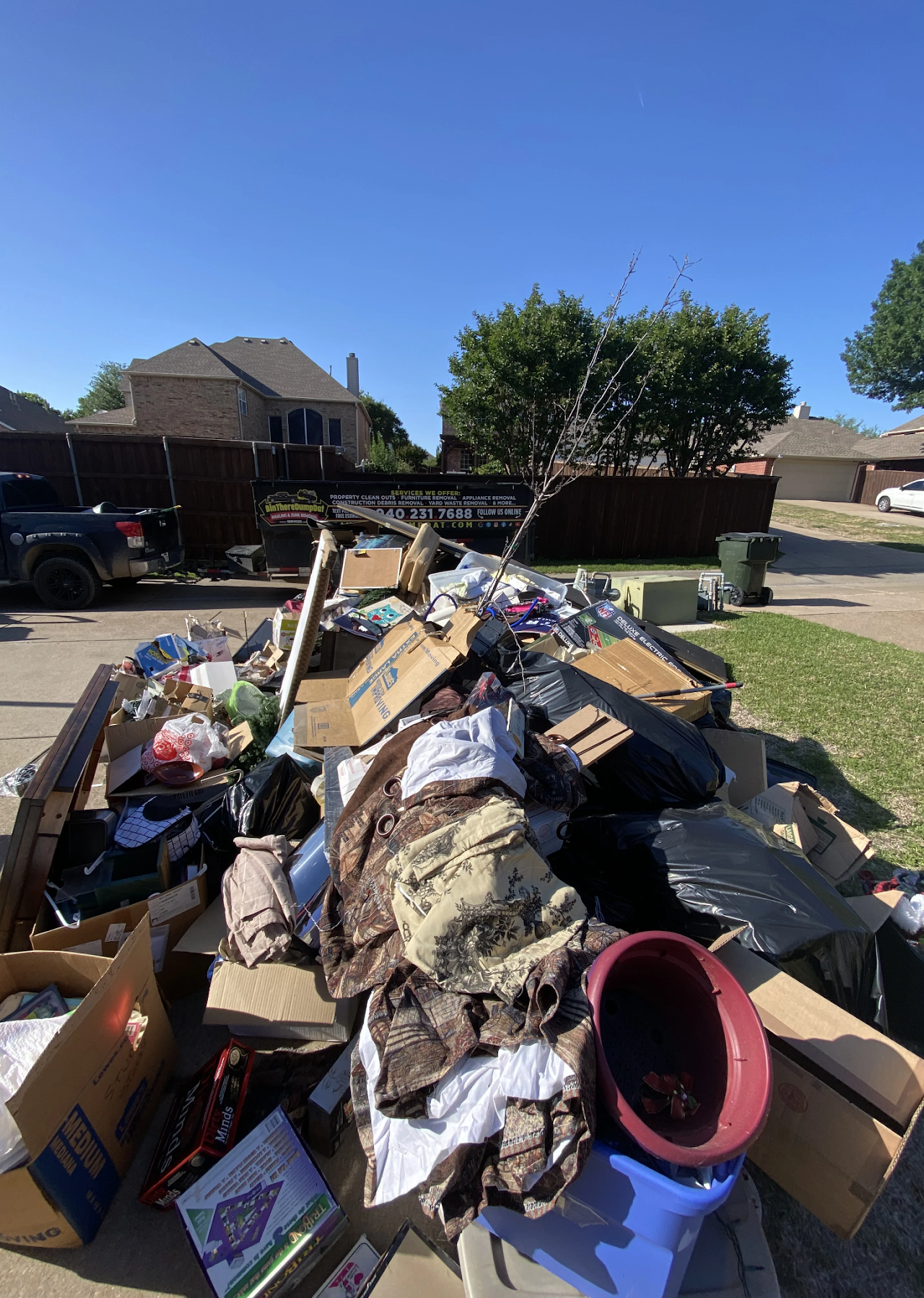 A large pile of clutter and junk, including boxes, trash bags, and household items, outside a house with a junk removal truck nearby.